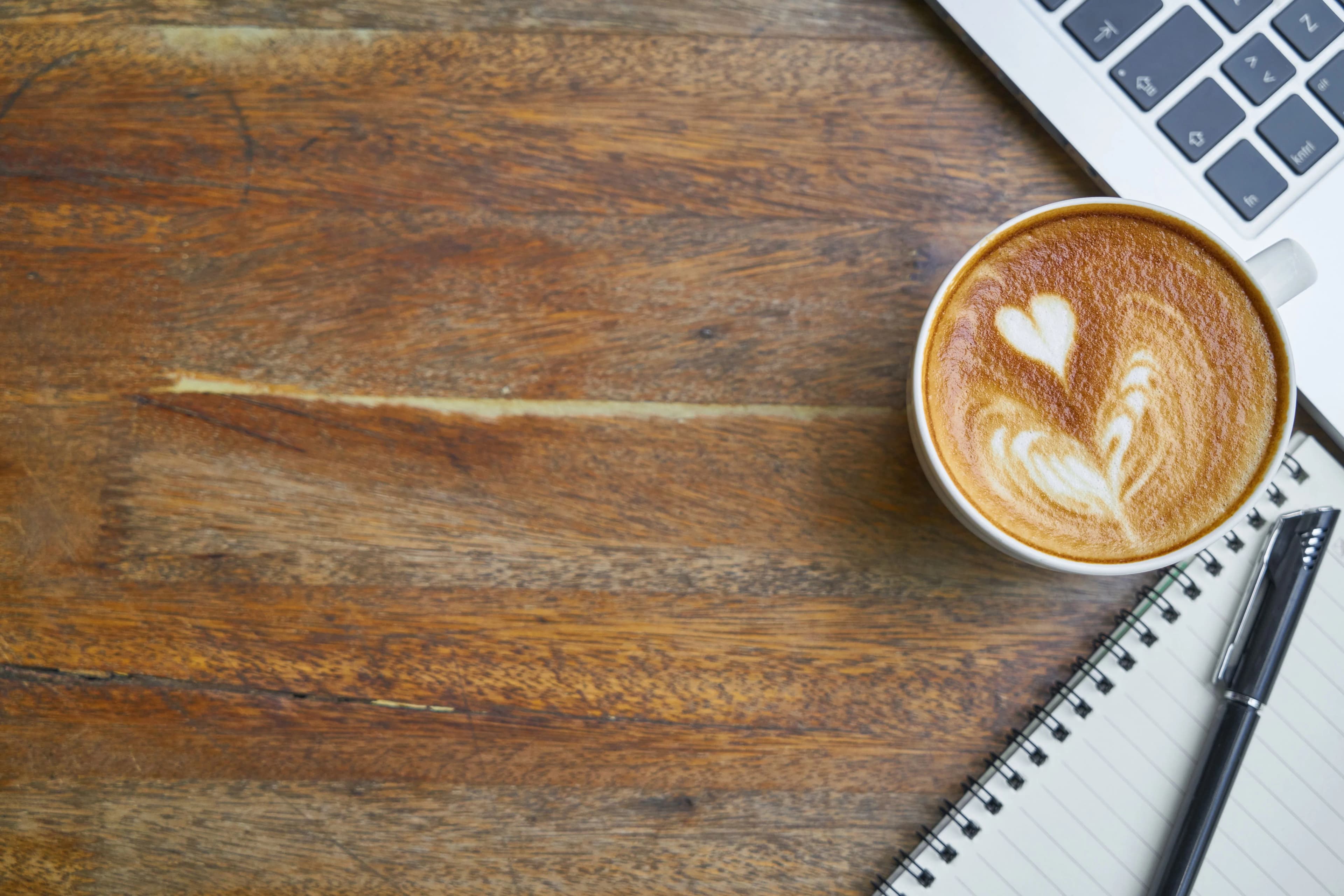 Secangkir cappuccino di dekat keyboard laptop dan notebook pada meja kerja bernuansa hangat
